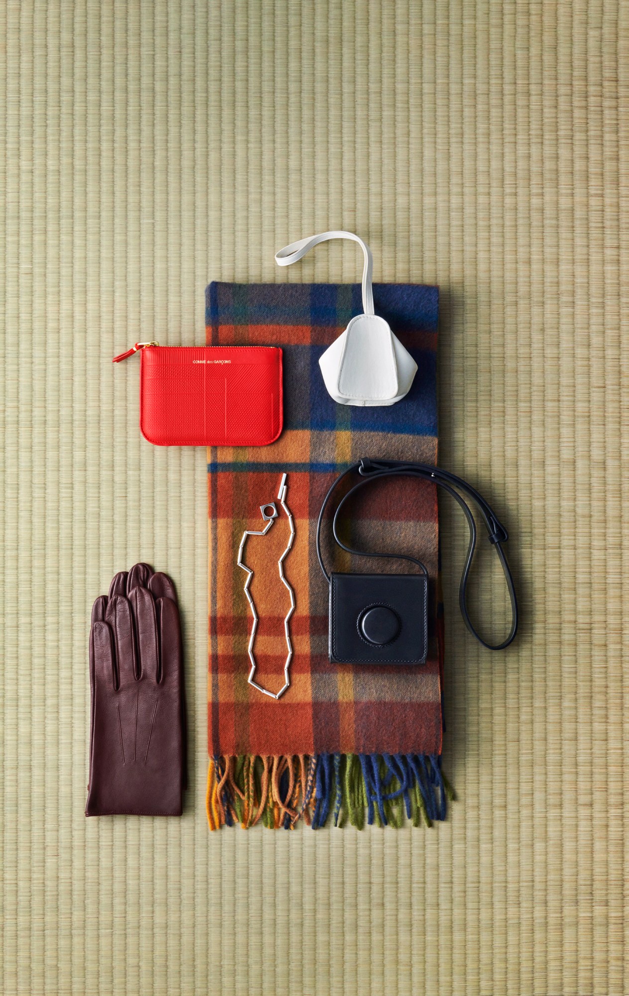 Flat lay of luxury accessories on woven mat including red wallet, plaid scarf, white bag, brown gloves and necklace