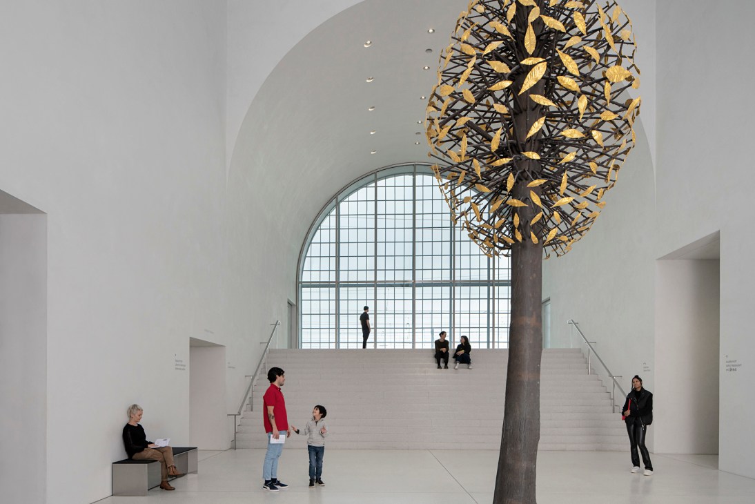 Modern art gallery interior with large sculptural tree installation and visitors on white floor