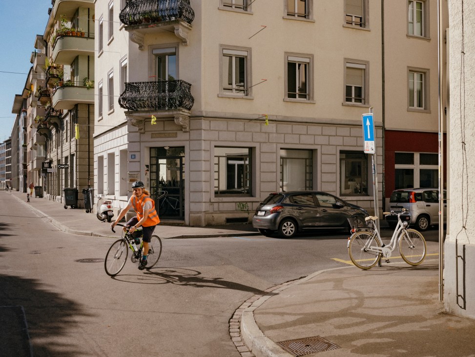Worker in high-visibility vest cycles through Zurich street past parked car and white bicycle