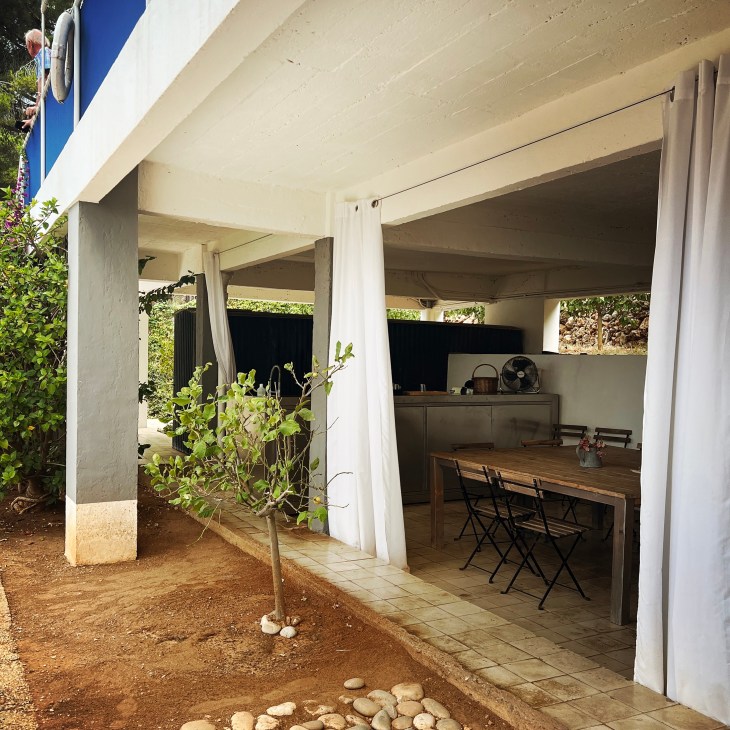 Modern outdoor kitchen with concrete structure, white curtains, wooden dining table and chairs under covered terrace