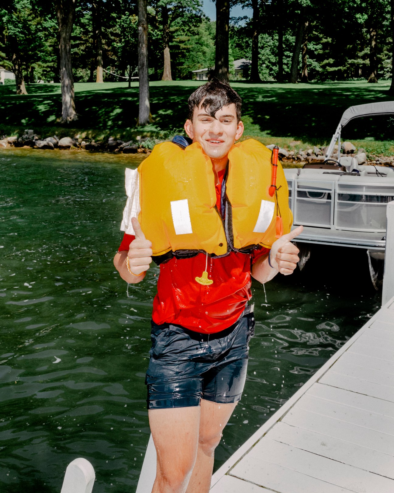 Young man in yellow life jacket standing waist-deep in lake water giving thumbs up near boat dock