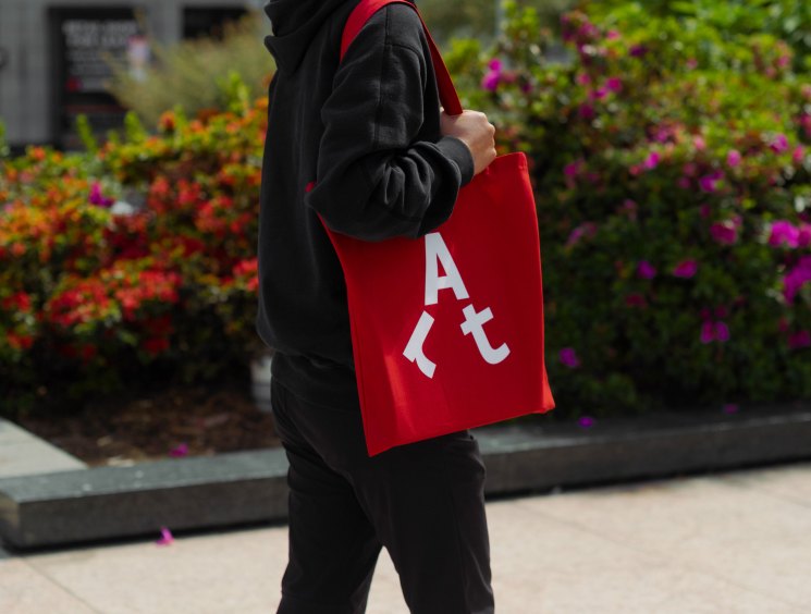 Person wearing black hoodie holding red tote bag with white "Art" lettering in front of colorful flower garden