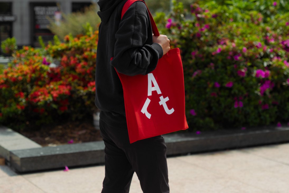 Person wearing black hoodie holding red tote bag with white "Art" lettering in front of colorful flower garden