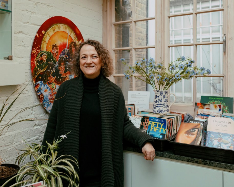 Geraldine Cox standing in Kirkdale Bookshop wearing green sweater with books and decorative plate visible