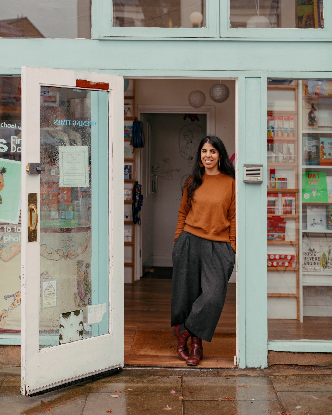 Portrait of Meera Ghanshamdas standing in doorway of Moon Lane Books wearing orange sweater and grey pants