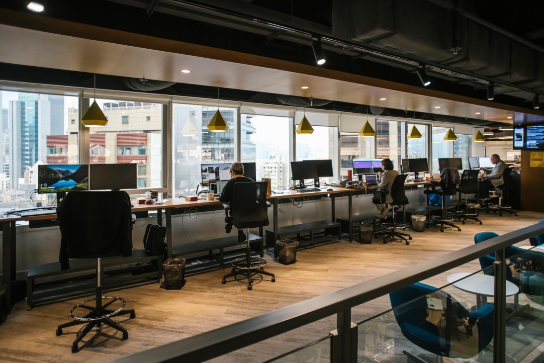 Modern office workspace with people at desks facing large windows overlooking urban cityscape