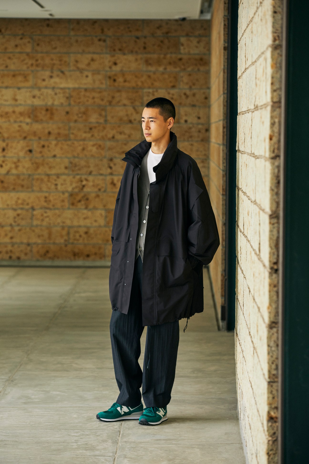 Young Asian man in dark coat and trousers standing against concrete wall in modern architectural space