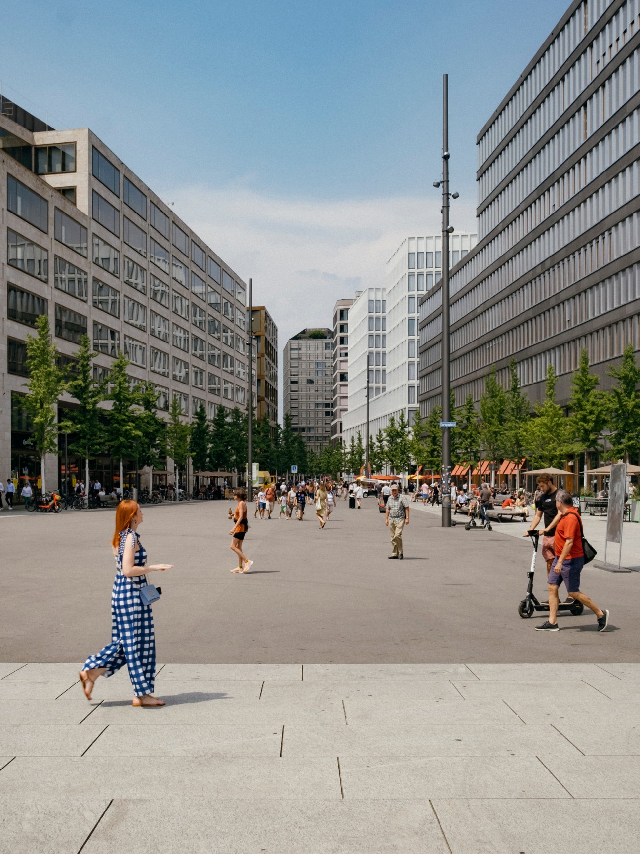 People walking and gathering in a modern urban plaza surrounded by contemporary office buildings and green trees