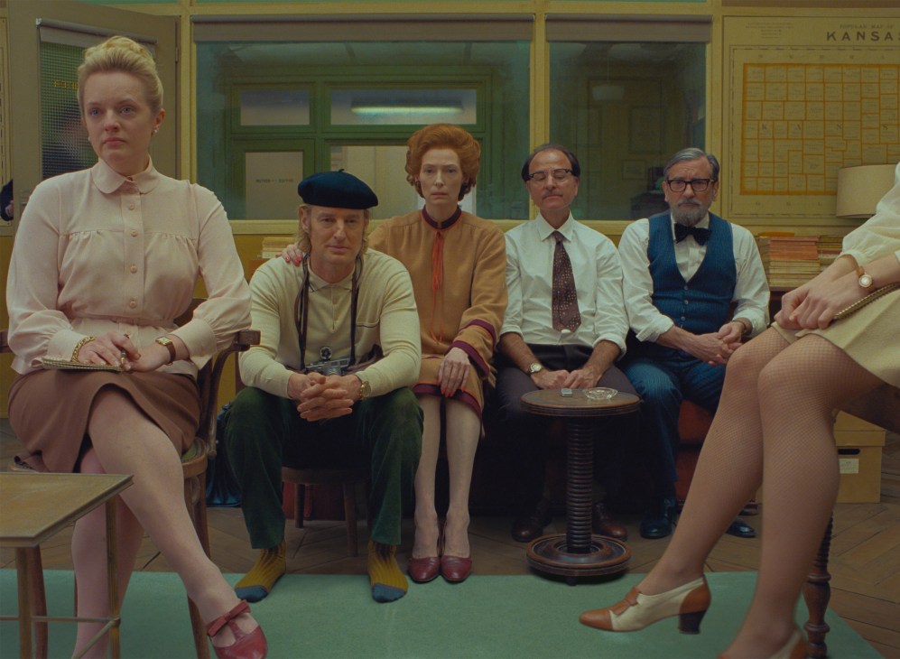 Six people in vintage clothing sitting in a row on benches in a retro-styled interior