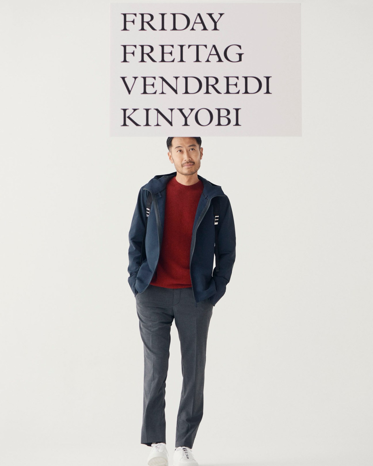 Model wearing black technical jacket, red jumper, grey trousers and white trainers against neutral background