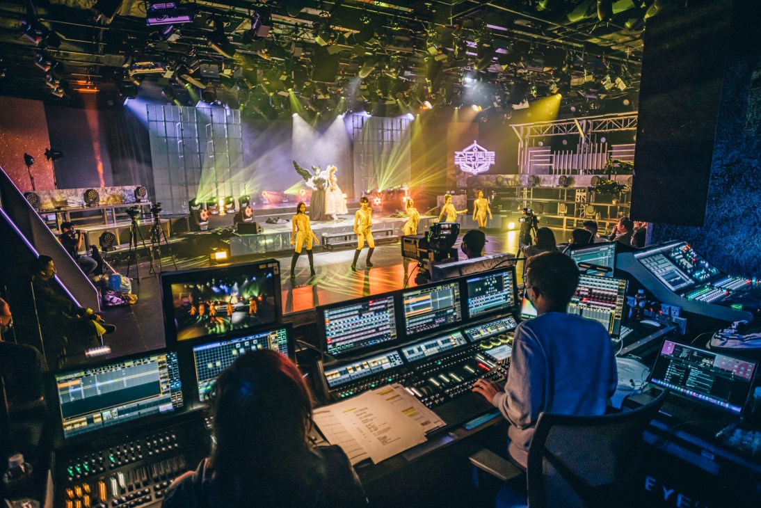 Television production control room with operator at mixing console overlooking illuminated studio stage with performers