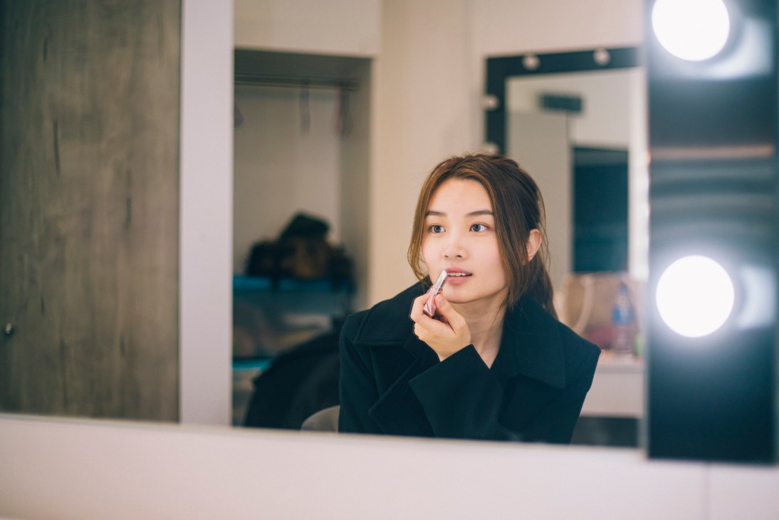 Young woman applying lipstick while looking in a mirror with professional lighting in background