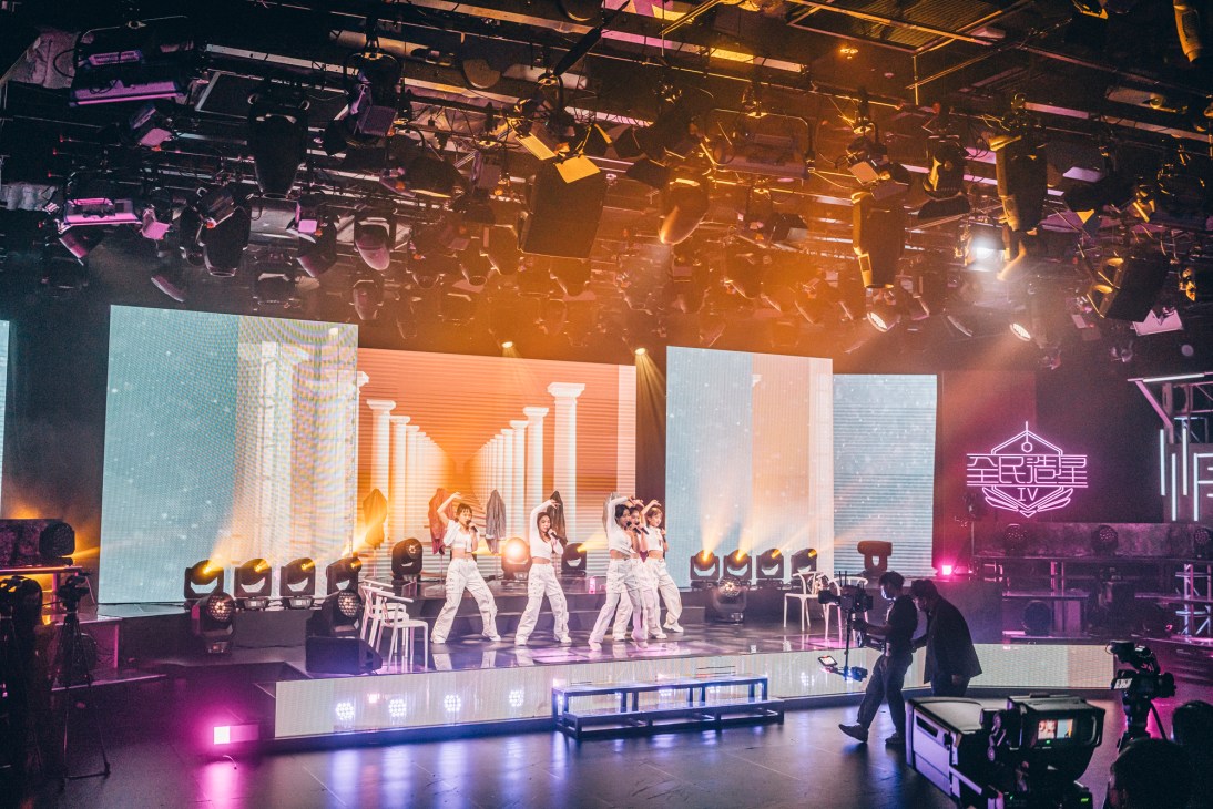 Female contestants in white outfits rehearsing dance moves on illuminated stage for King Maker TV show