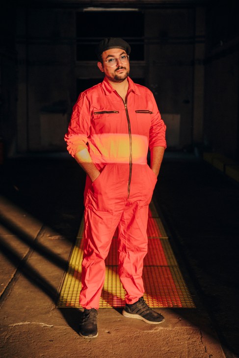 Michel Niknafs wearing red overalls and a black beret stands on a lit floor in a warehouse venue.
