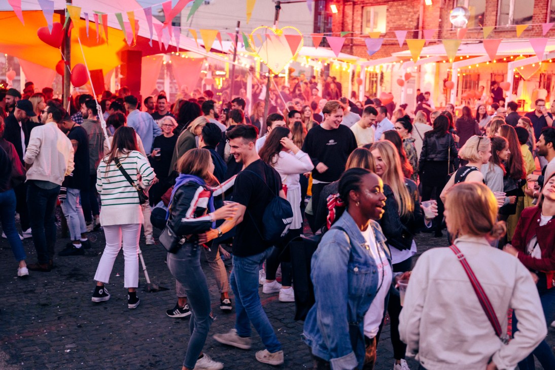 Large crowd of people dancing and socializing in a colorful lit courtyard with bunting decorations at Nåpoleon Komplex.