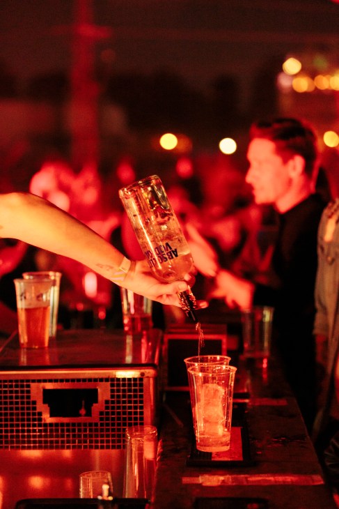 Bartender pouring a drink at a nightclub bar with red lighting and blurred revellers in background.