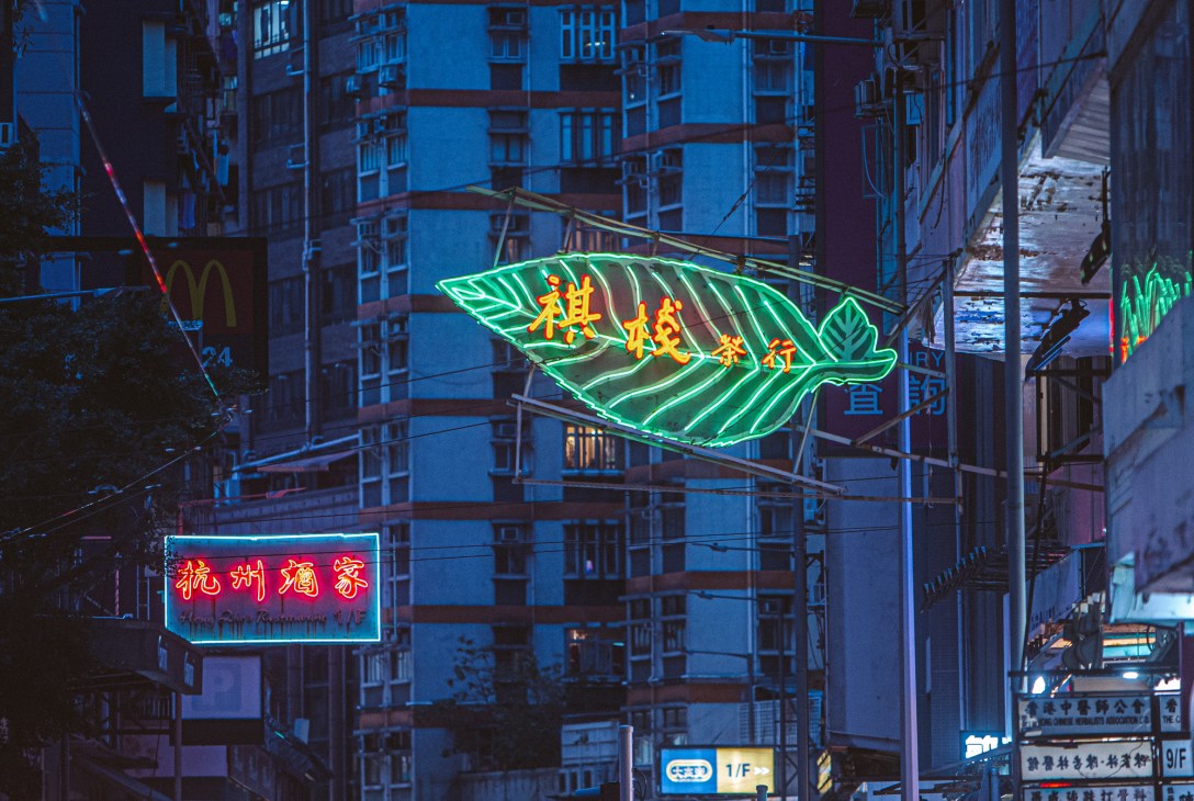Neon sign in a narrow Hong Kong alley with green leaf design and Chinese characters, plus red restaurant sign below