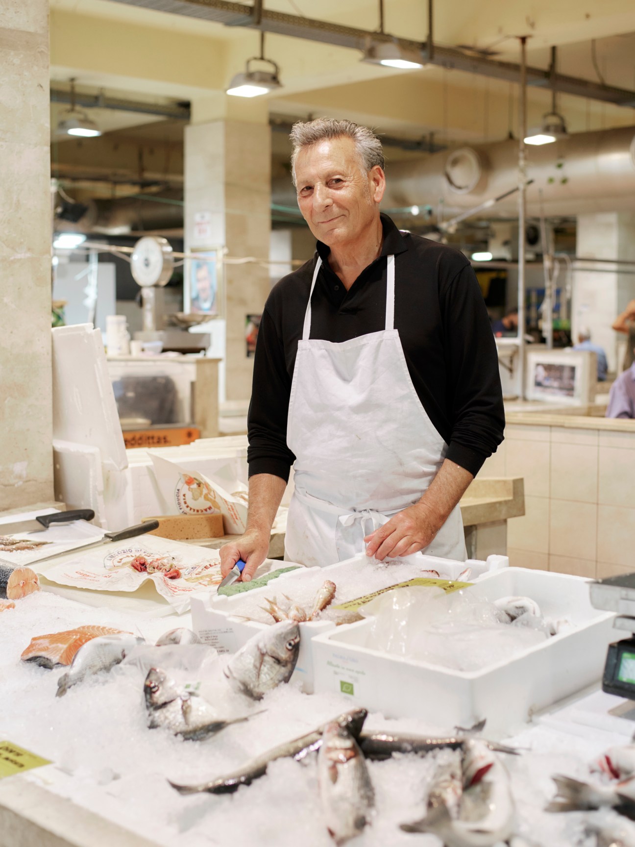 Smiling fishmonger in white apron stands behind display of fresh fish at indoor market counter