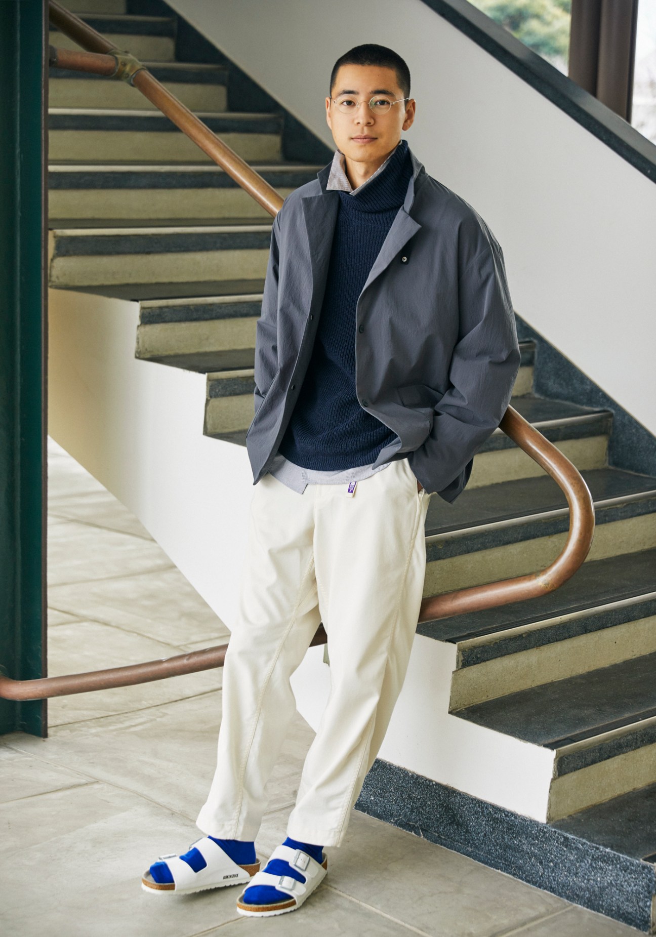 Man in gray jacket and navy sweater standing by modern staircase wearing Birkenstock sandals