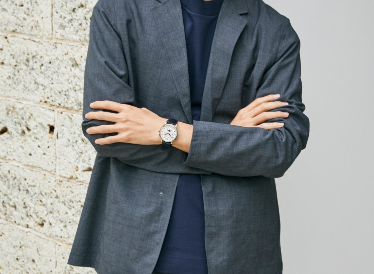Asian man in grey suit jacket and navy t-shirt with crossed arms wearing watch against textured wall