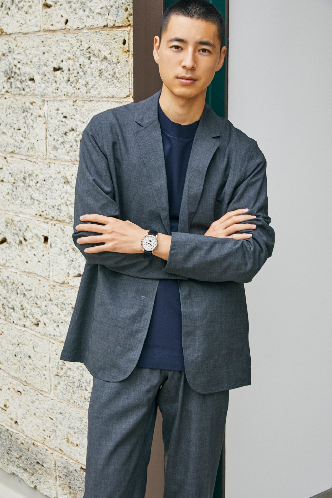 Asian man in grey suit jacket and navy t-shirt with crossed arms wearing watch against textured wall