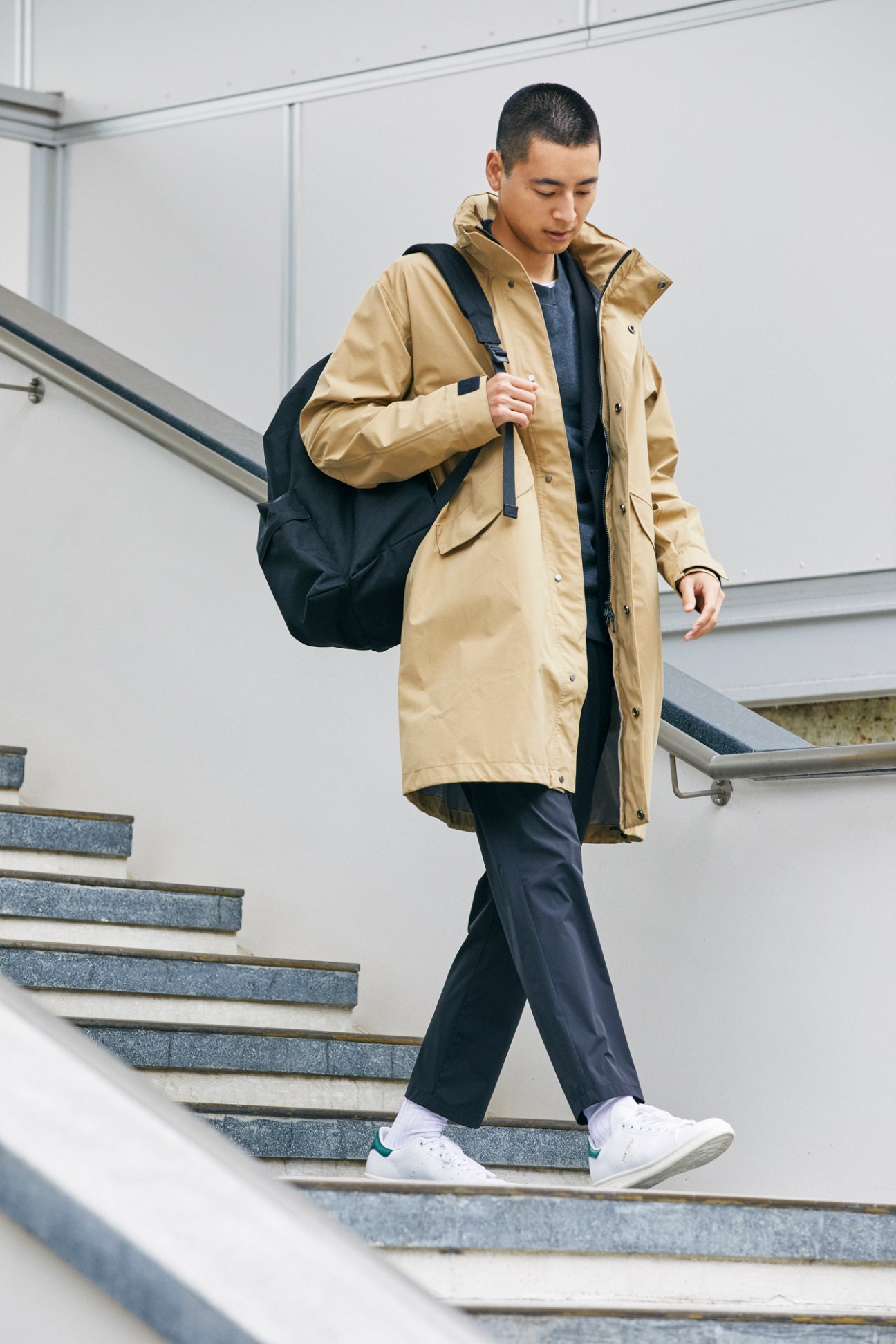 Asian man walking down stairs wearing tan coat, dark pants, white sneakers and carrying black backpack