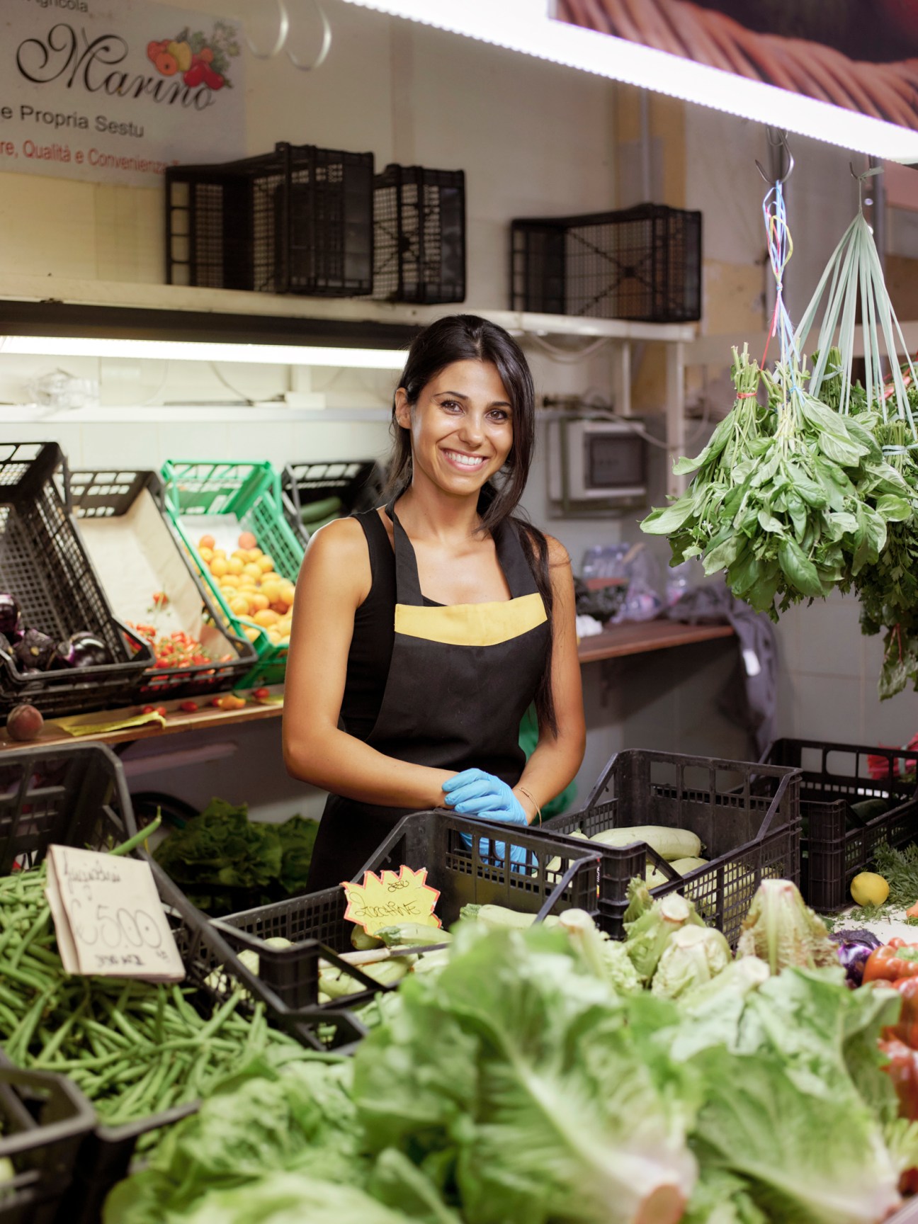 Smiling woman in black apron arranging fresh leafy vegetables at San Benedetto Market produce stand