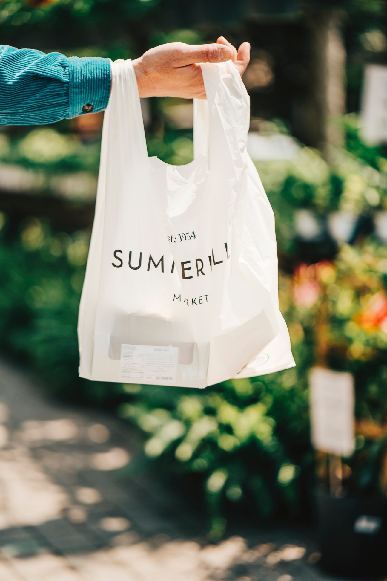 Hand holding white plastic shopping bag with Summerhill Market logo and est. 1954 text against blurred green background