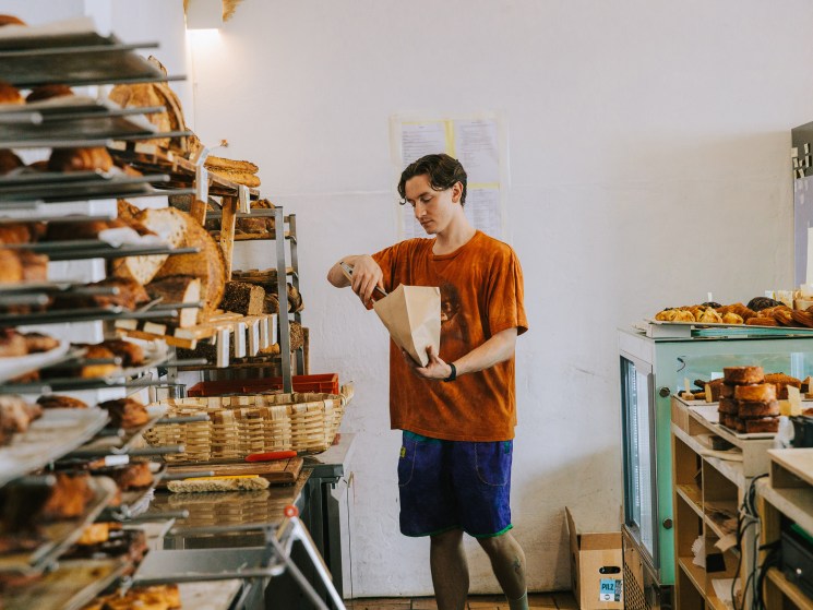 Baker in orange shirt packaging pastries into paper bag at La Maison bakery with bread shelves and display case