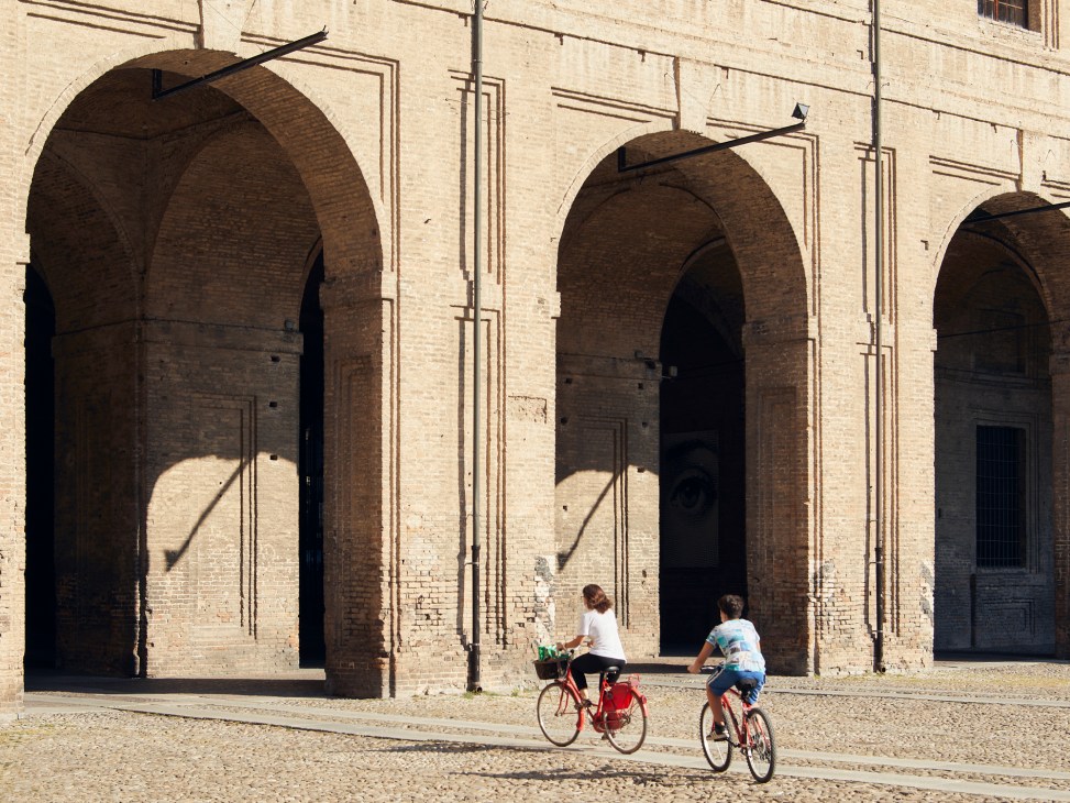 Two cyclists riding past historic arched colonnade architecture in Parma, Italy