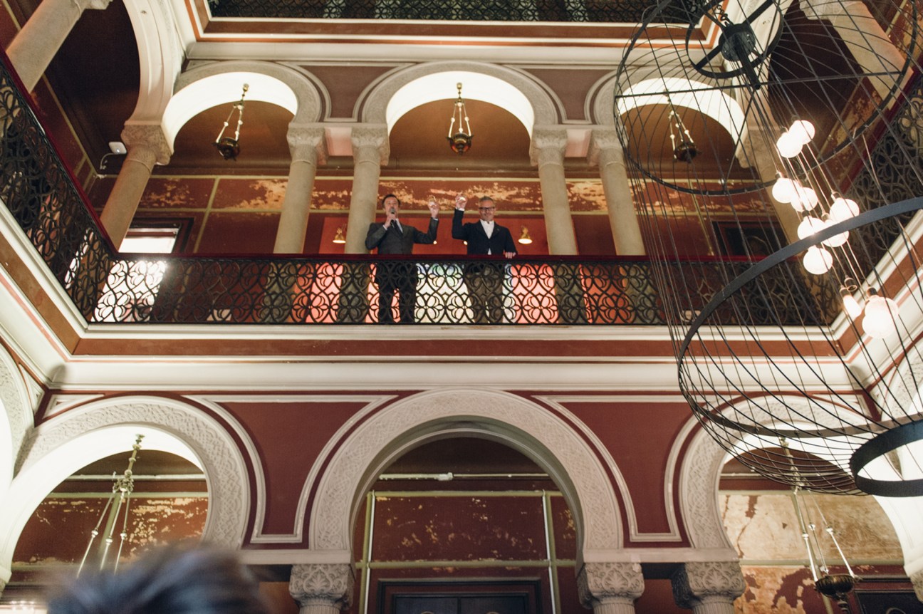 Two men standing on ornate balcony with decorative ironwork and classical columns in grand interior space
