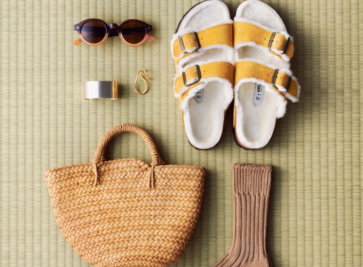 Flat lay of accessories on woven mat: sunglasses, yellow sandals, jewelry, woven bag, and ribbed socks