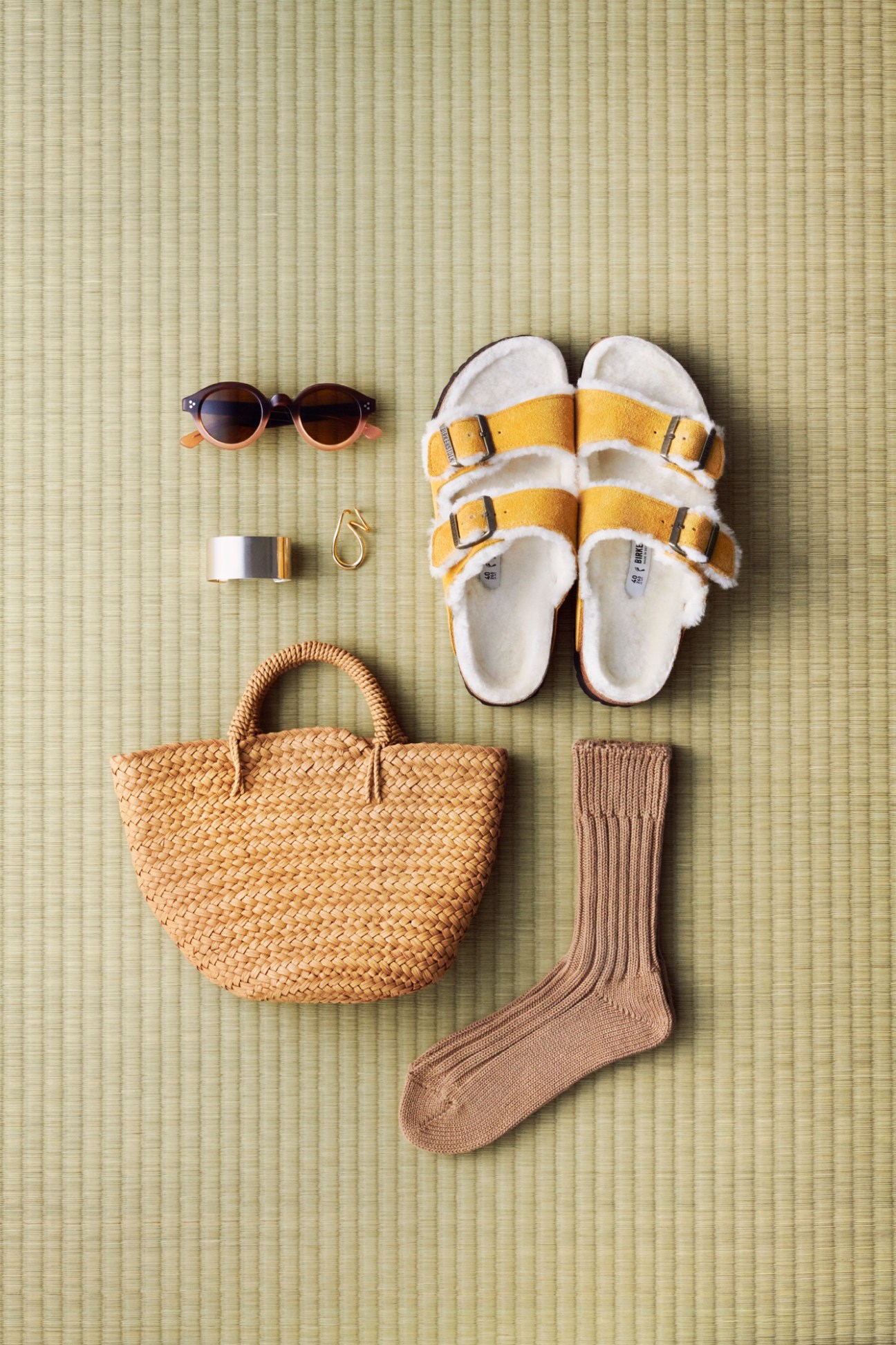 Flat lay of accessories on woven mat: sunglasses, yellow sandals, jewelry, woven bag, and ribbed socks