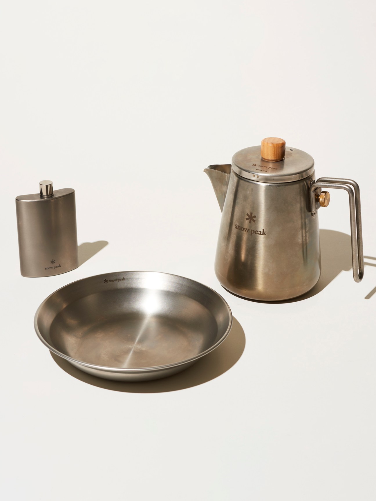Stainless steel kettle with wooden knob, metal flask, and shallow plate arranged on light background