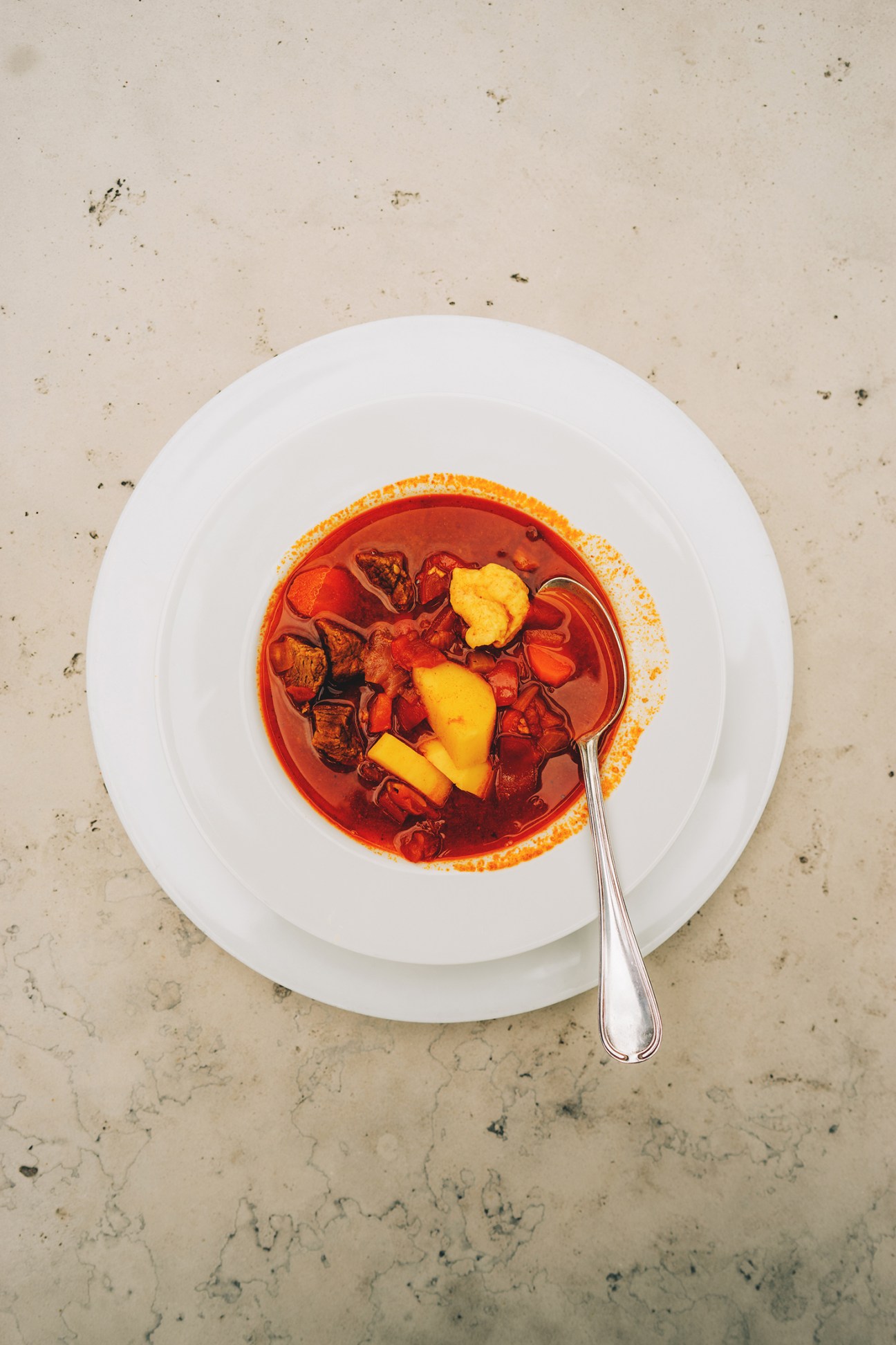 A bowl of kettle goulash with vegetables and meat in rich red broth, served in a white bowl with a spoon