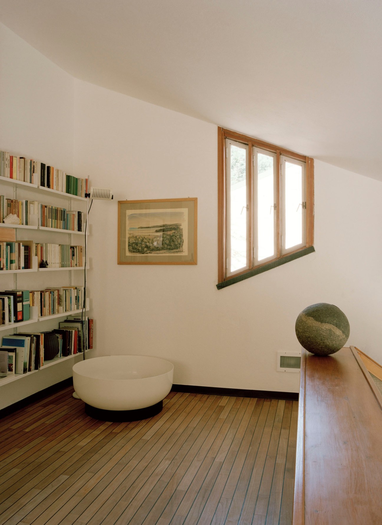 Modern reading nook with white built-in bookshelves, round white ottoman, wooden bench and three tall windows