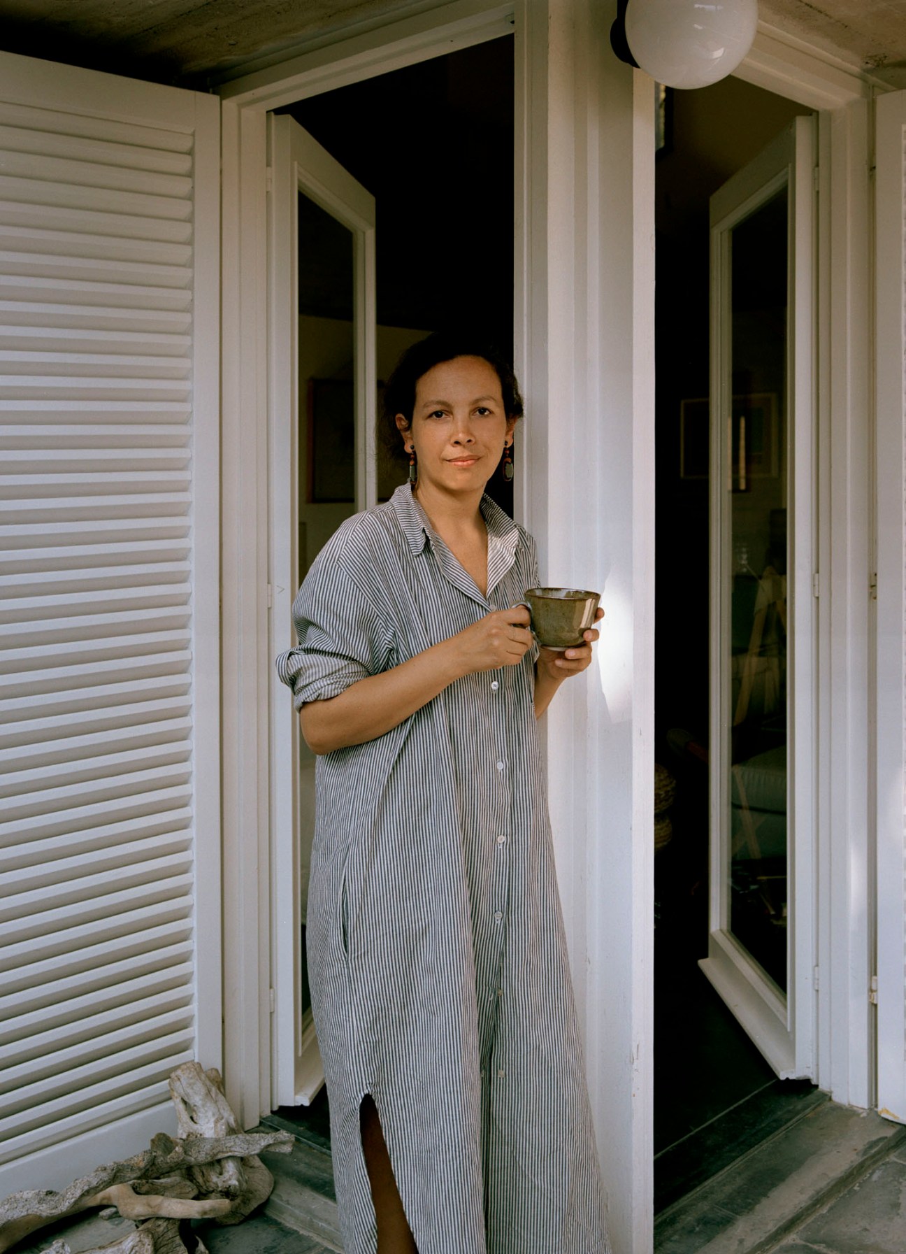 Woman in striped shirt dress holding ceramic mug standing in doorway of white building