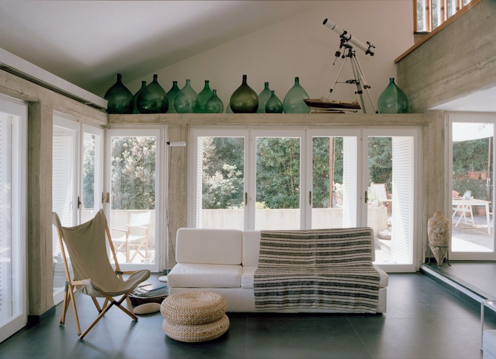 Modern living room with white sofa, striped throw, folding chair, telescope, and green glass bottles on shelf above windows