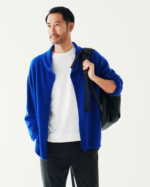 Man wearing blue cardigan, white t-shirt, black trousers and black backpack against white background.