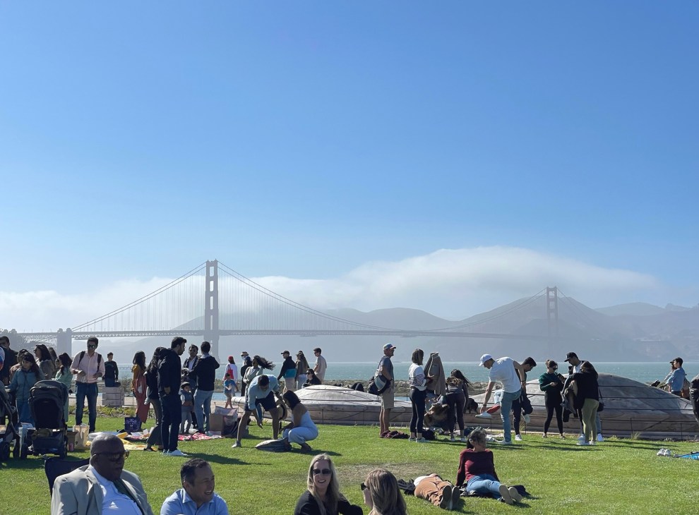 People sitting and standing on grassy area with Golden Gate Bridge visible in background on clear day