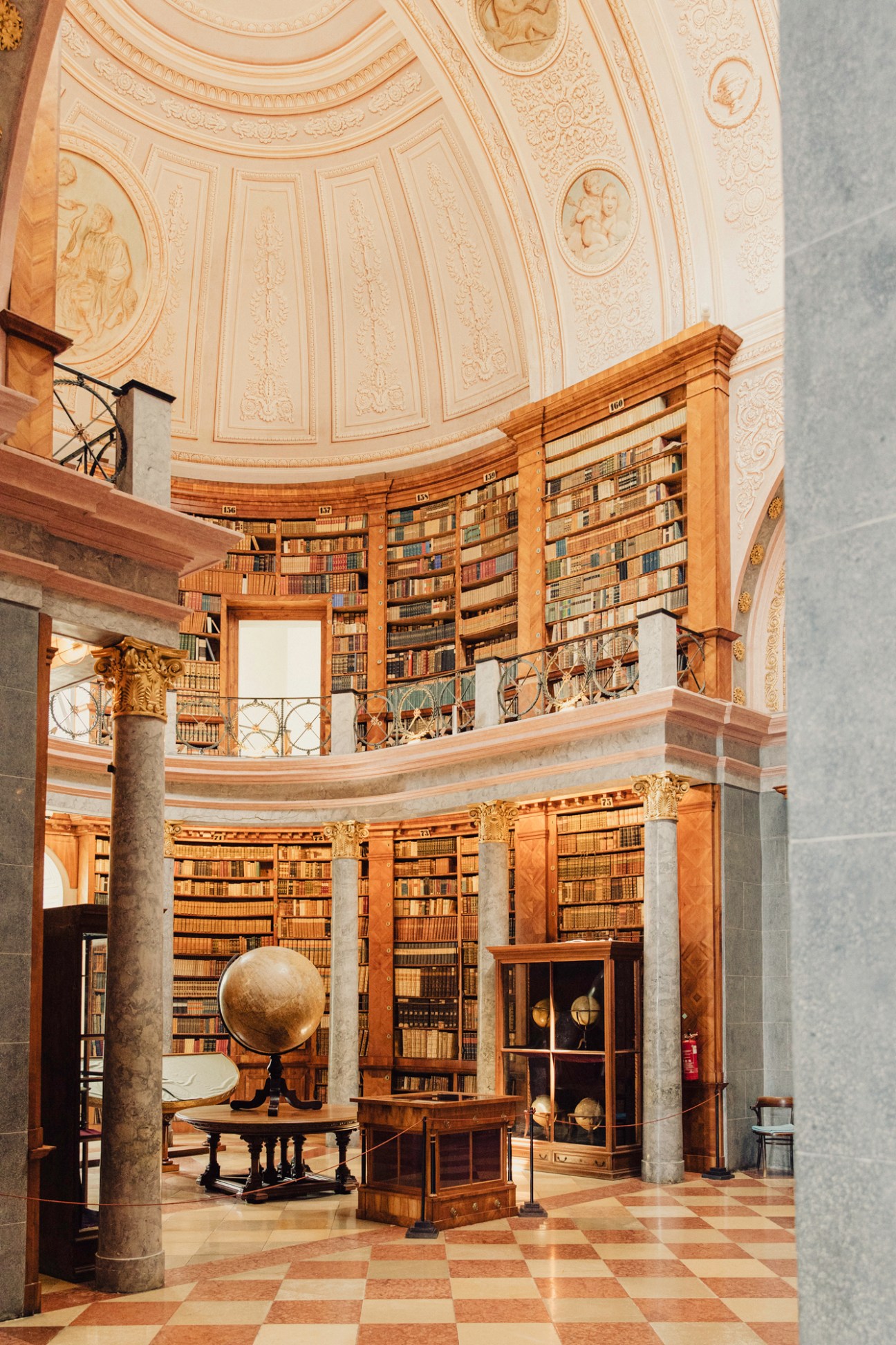Grand circular library interior with ornate gilded ceiling, marble columns, and floor-to-ceiling bookshelves on two levels