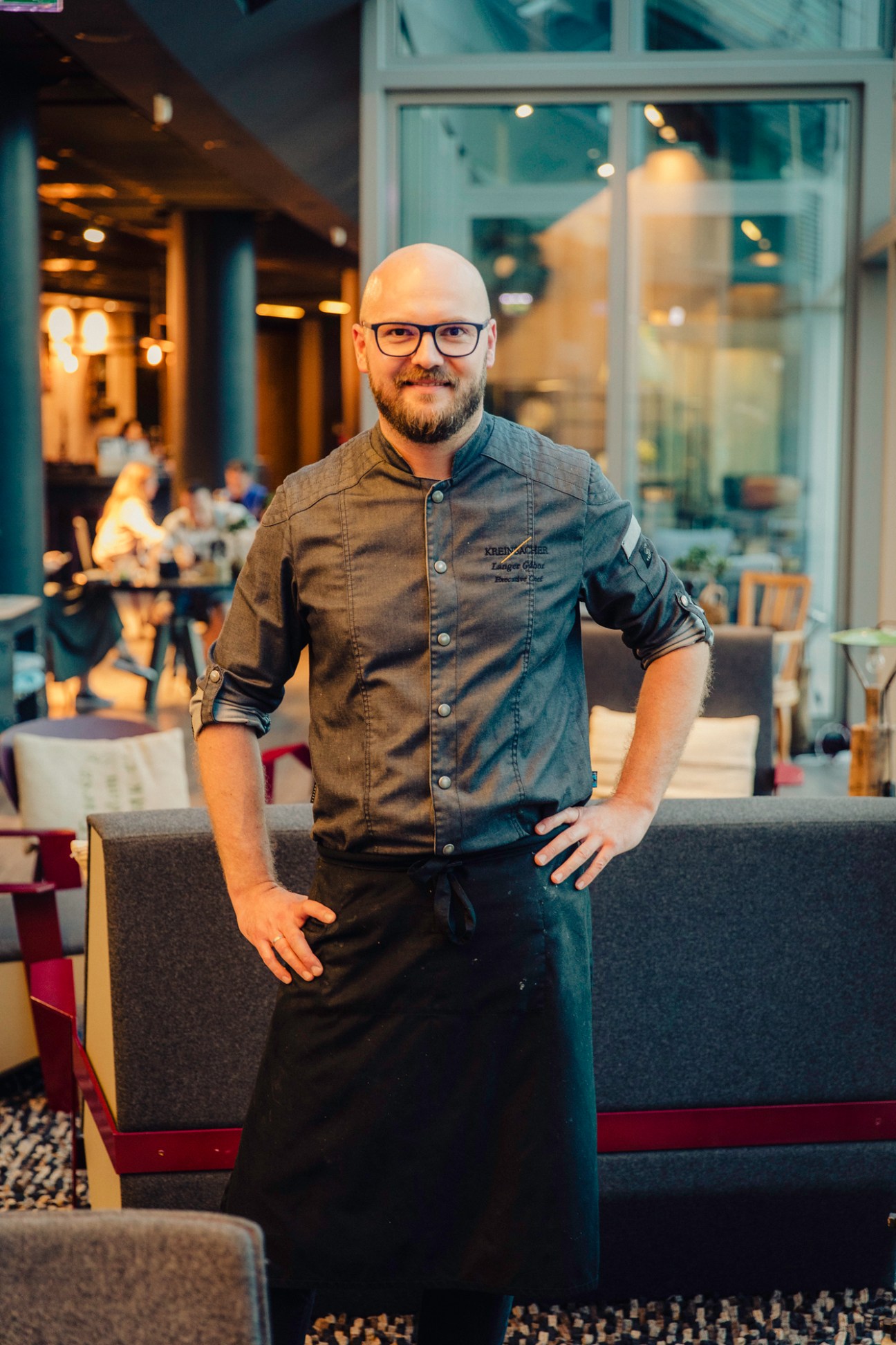 Bald man with glasses and beard in chef's coat standing in modern restaurant interior