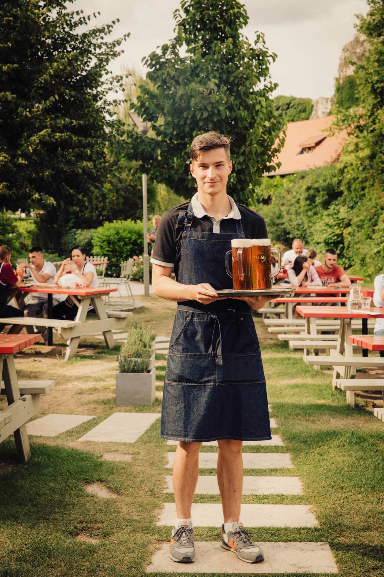 Young waiter in denim apron carrying beer mug on tray at outdoor beer garden