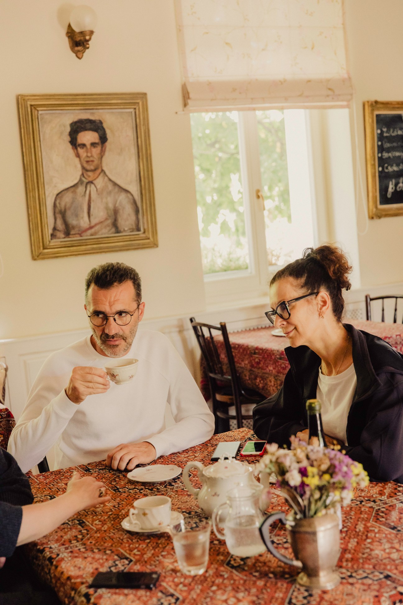 Two people having tea at a table in an elegant room with framed portraits and traditional decor