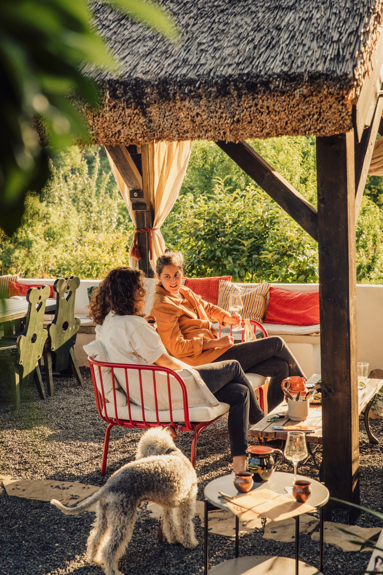 Two women relaxing on outdoor terrace with thatched roof pergola, small white dog, and drinks on table