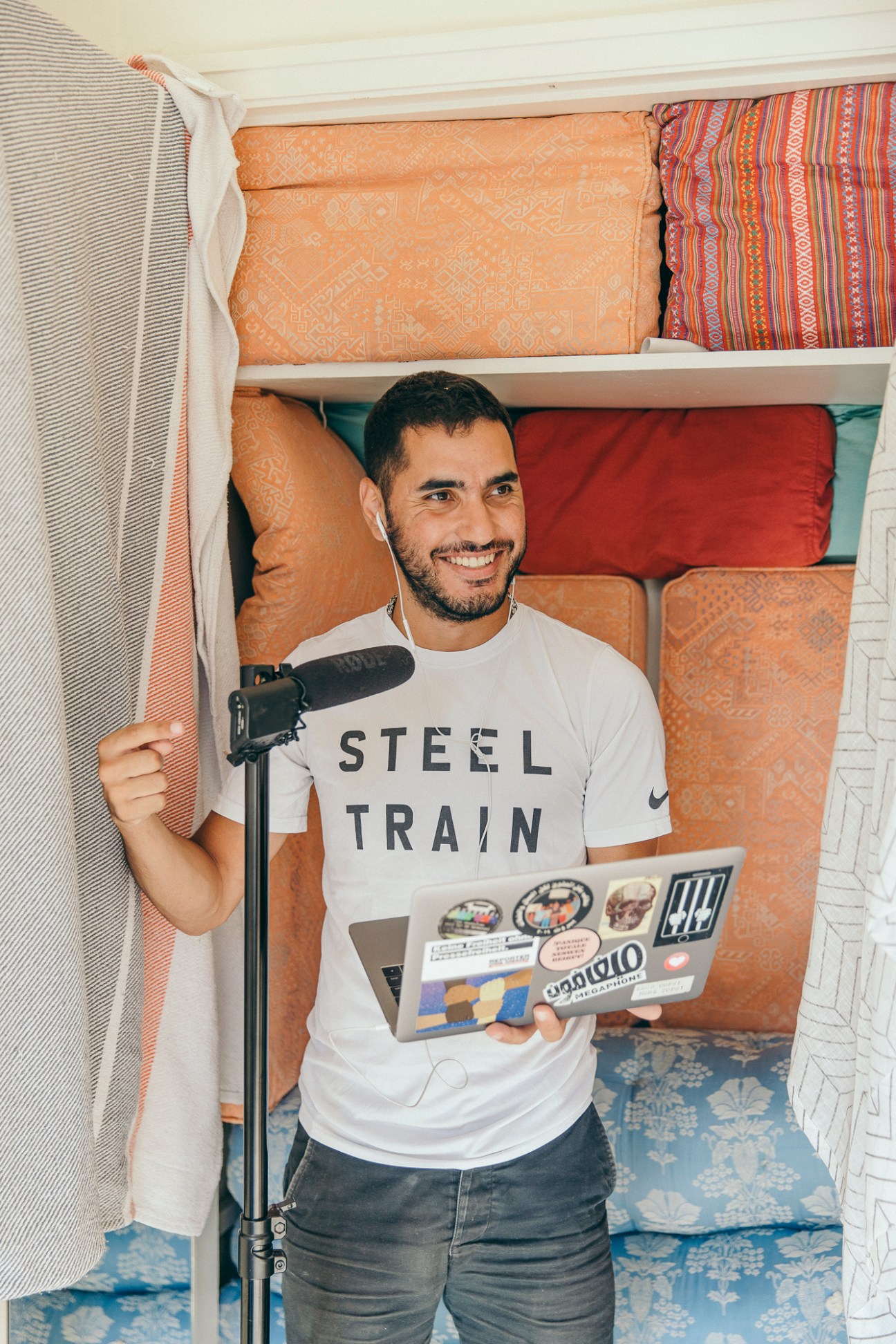 Man in white Steel Train t-shirt recording podcast with microphone and laptop in colorful van interior