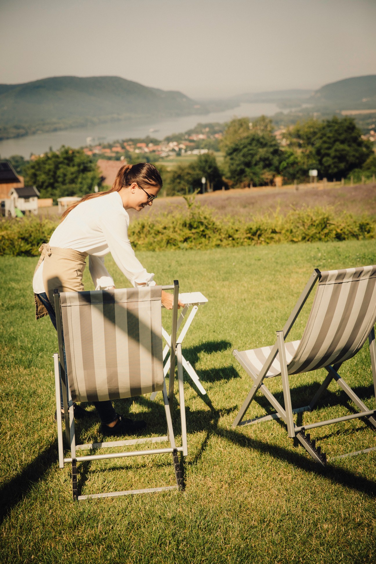 Woman setting up outdoor dining table with striped deck chairs on grassy hillside overlooking mountains