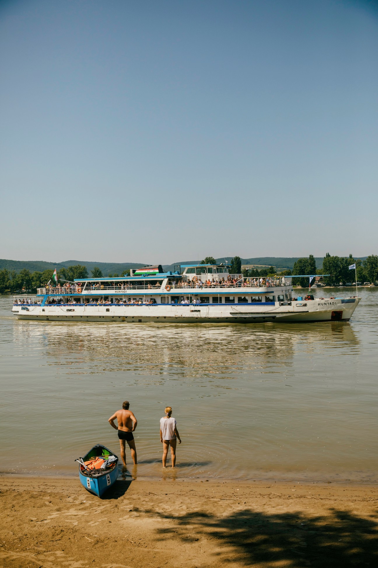 Two swimmers in shallow Danube water watching a passenger ferry boat full of tourists.