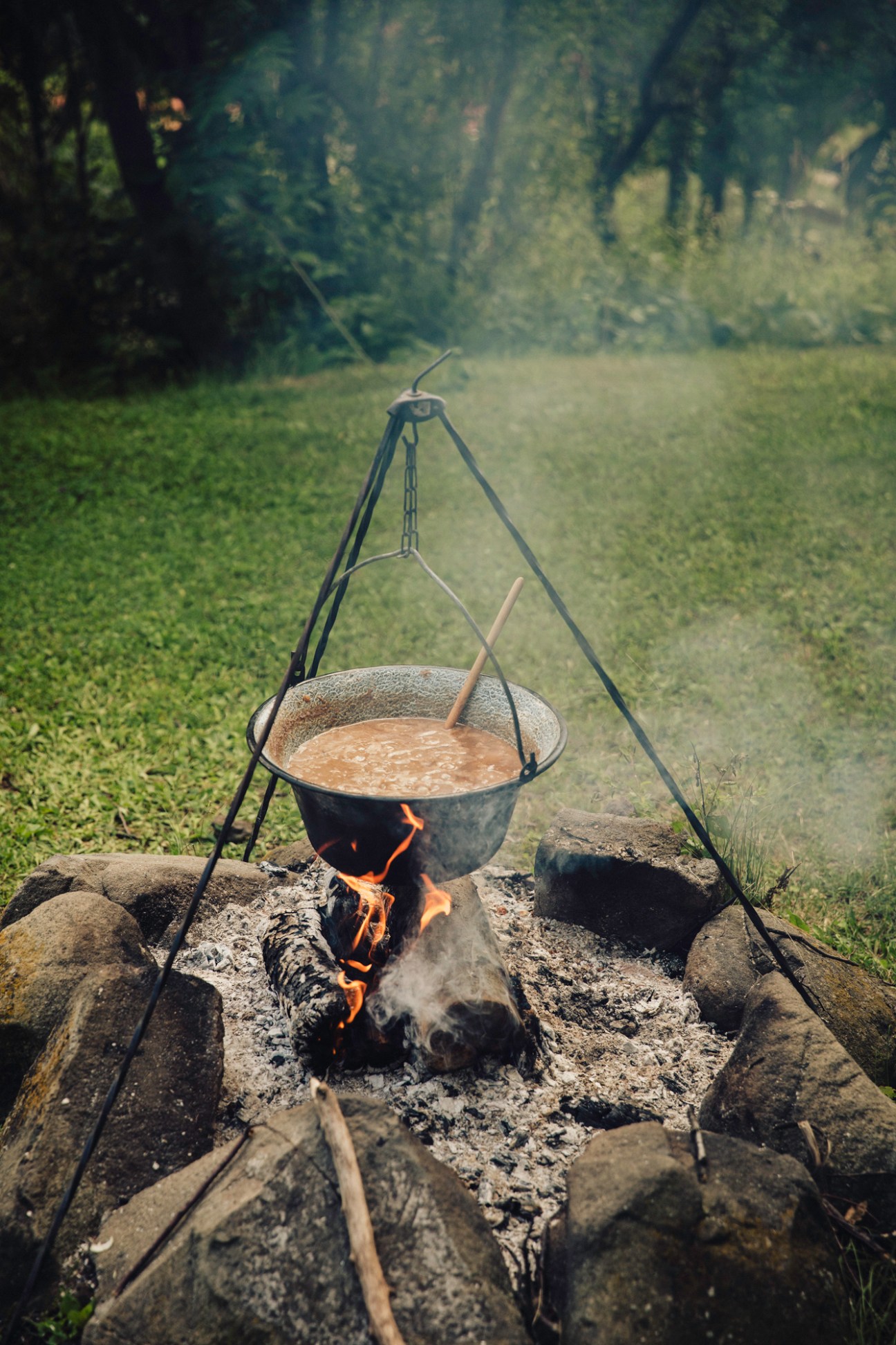Outdoor campfire cooking with cast iron pot hanging from tripod over flames in forest clearing