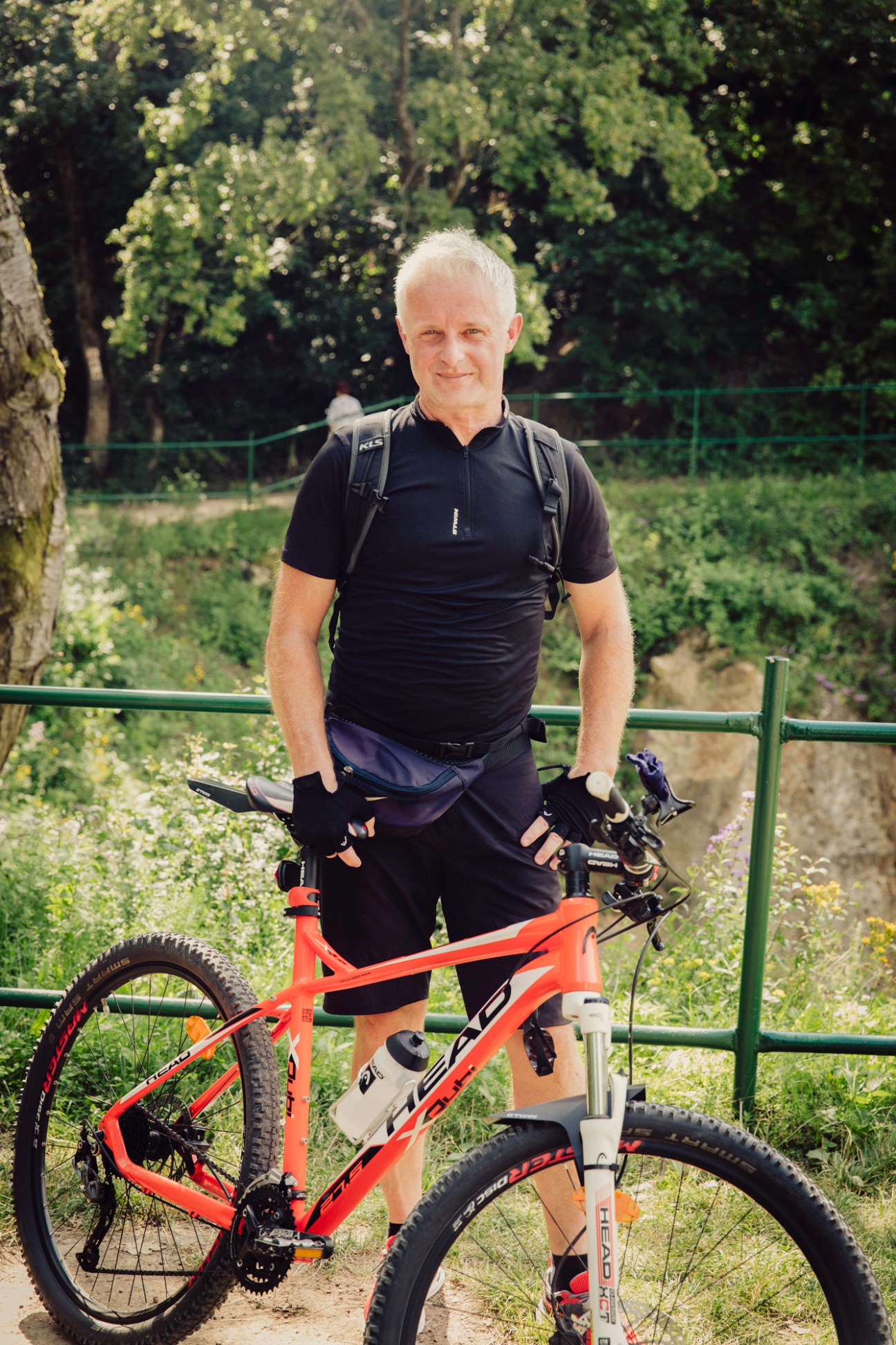 Man with gray hair standing behind orange mountain bike in outdoor park setting